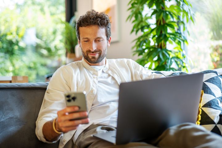 Muž sedí na gauči s laptopom a mobilným telefóonom v ruke